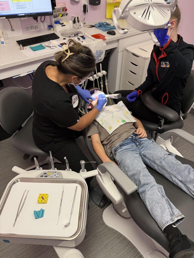A child reclines in a dental chair while two professionals perform treatment, one working on the mouth and the other assisting. The clinical setup includes a tray of dental tools, a counter with supplies, and a computer, emphasizing a modern pediatric care environment.